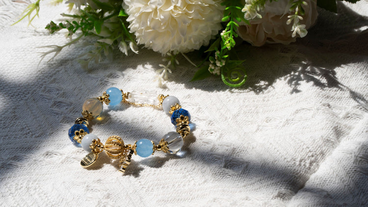 Handcrafted crystal bracelet featuring a blend of blue, clear, and gray beads accented with elegant gold charms and a decorative clasp, set on textured white fabric with green leaves and white flowers in the background.